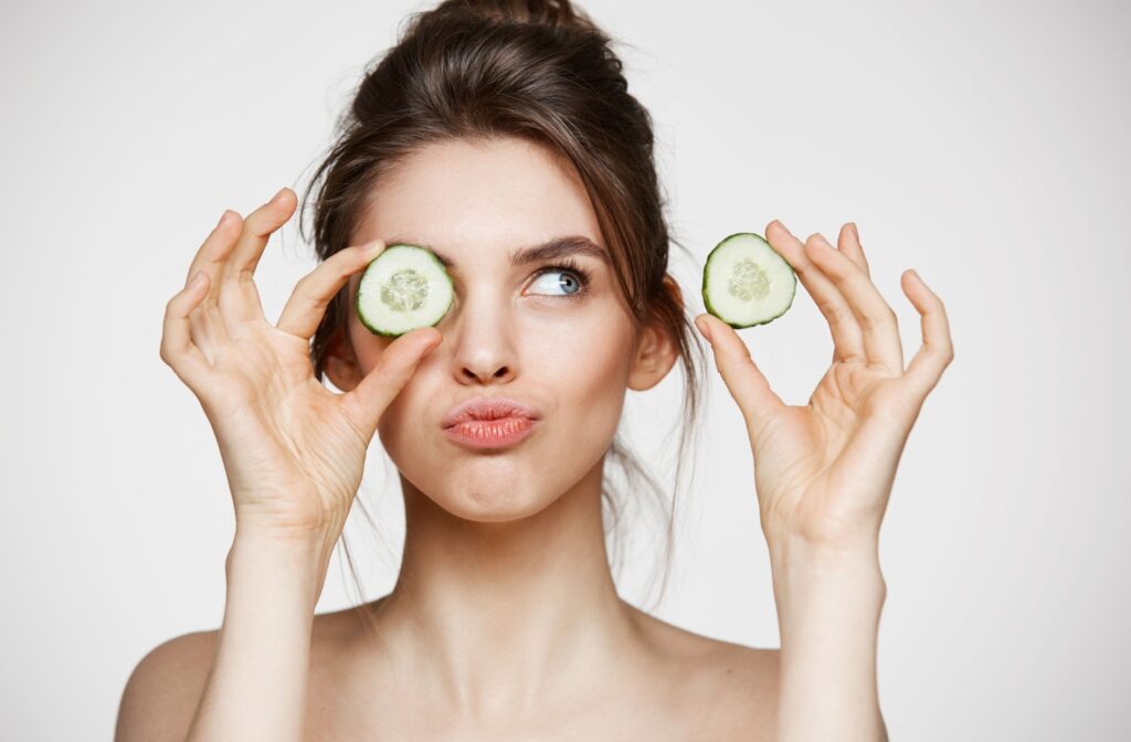 A person with their hair tied in a bun holds cucumber slices in front of their eyes for skin tightening