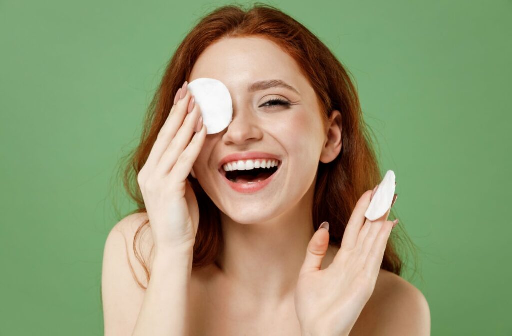 A person with a cheerful smile covers their left eye with a cotton round.