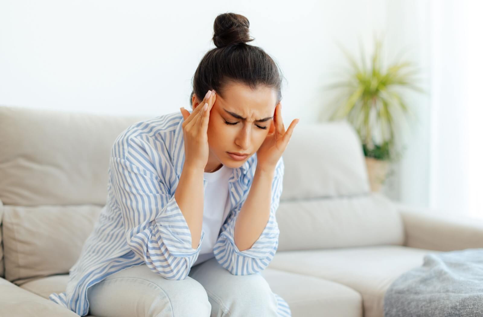 A person in a striped shirt sitting on a couch with eyes closed and hands on temples, illustrating how eye fatigue can cause dizziness and headaches.