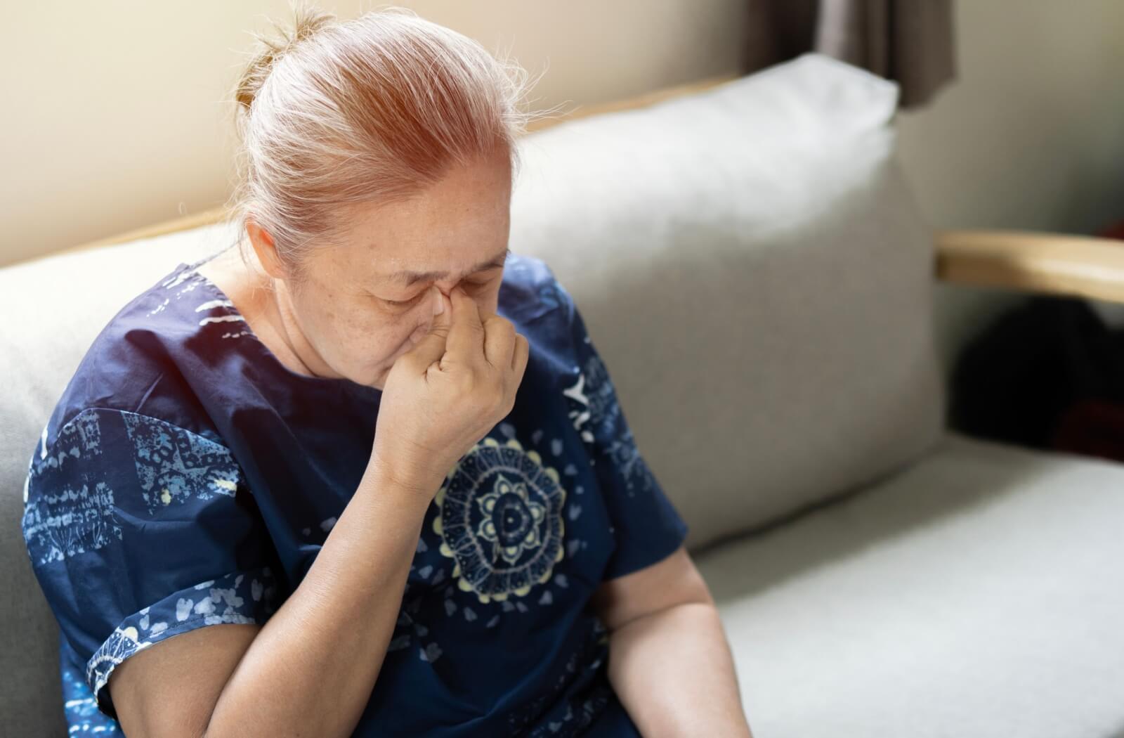 A person sitting on a sofa rubbing the bridge of their nose with eyes closed, showing symptoms of Meibomian Gland Dysfunction.