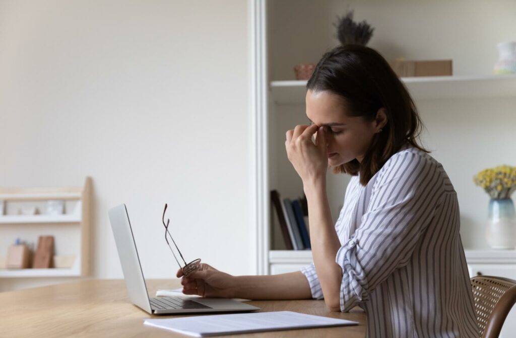A person sitting at a desk with a laptop, eyes closed and rubbing the bridge of their nose while holding a pair of eyeglasses, demonstrating eye strain or Meibomian Gland Dysfunction symptoms.