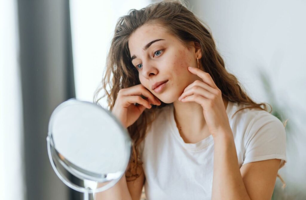 A young woman in a white t-shirt looking into a mirror and touching her face to examine red, irritated areas on her skin.